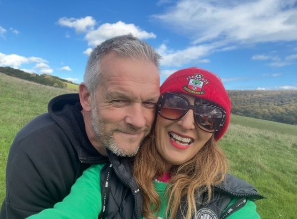 A women with long hair, sunglasses and a red woolly hat smiles at the camera next to a man with grey hair and black hoodie.